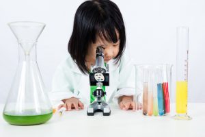 Junges Mädchen schaut, gekleidet in einem weißen Kittel, in eine Mikroskop.