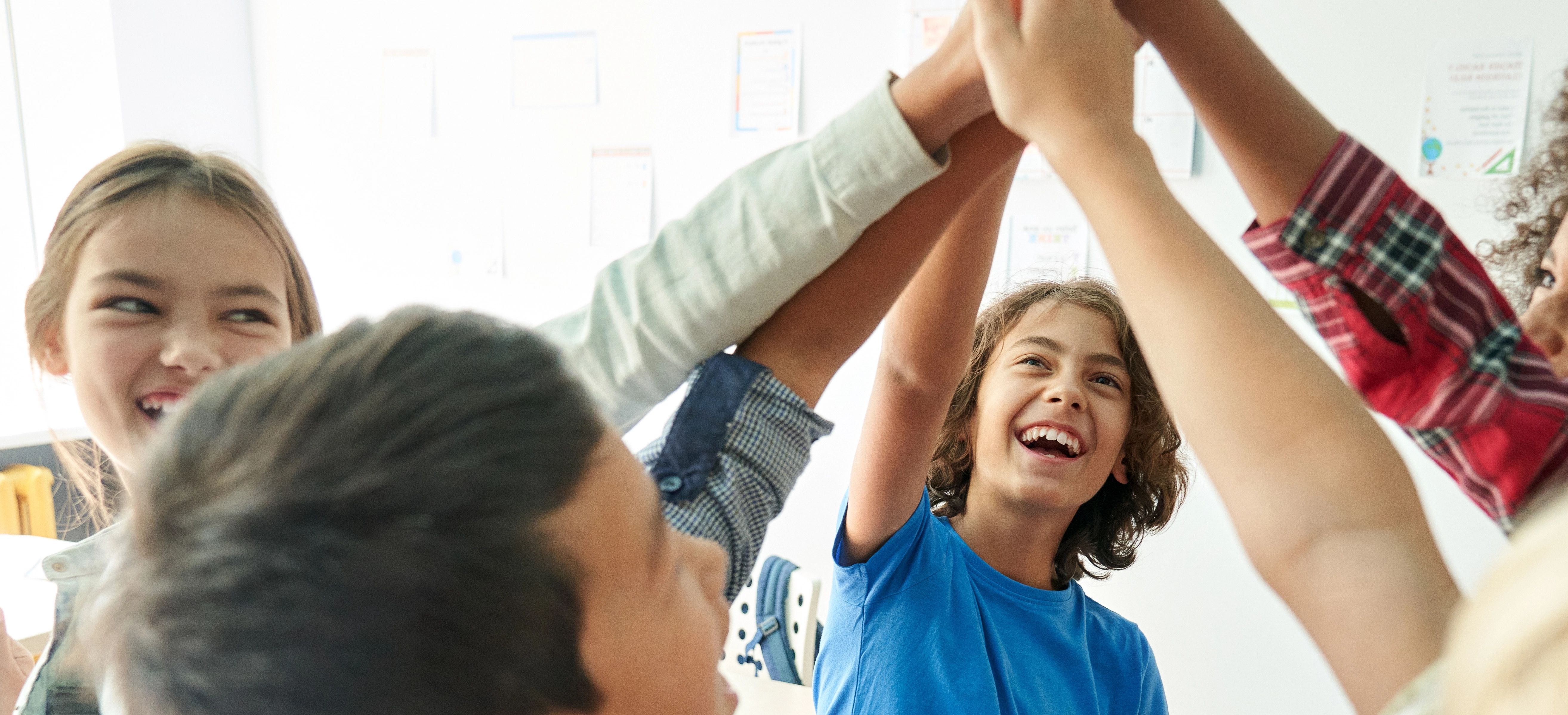 Drei junge Kinder in einem Klassenraum lehnen sich zusammen, um auf den Bilddschirm eines silberfarbenen Laptops zu schauen.