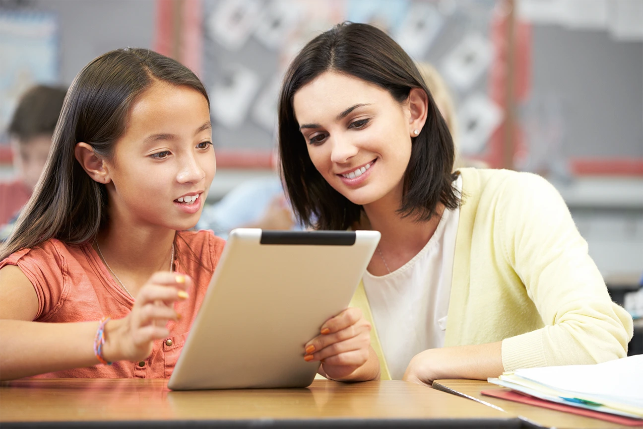 Eine Schülerin und eine Lehrerin sitzen zusammen in einem Klassenzimmer, lächeln und schauen auf ein Tablet. Der Schüler zeigt auf den Bildschirm, während der Lehrer ihm Anweisungen gibt.